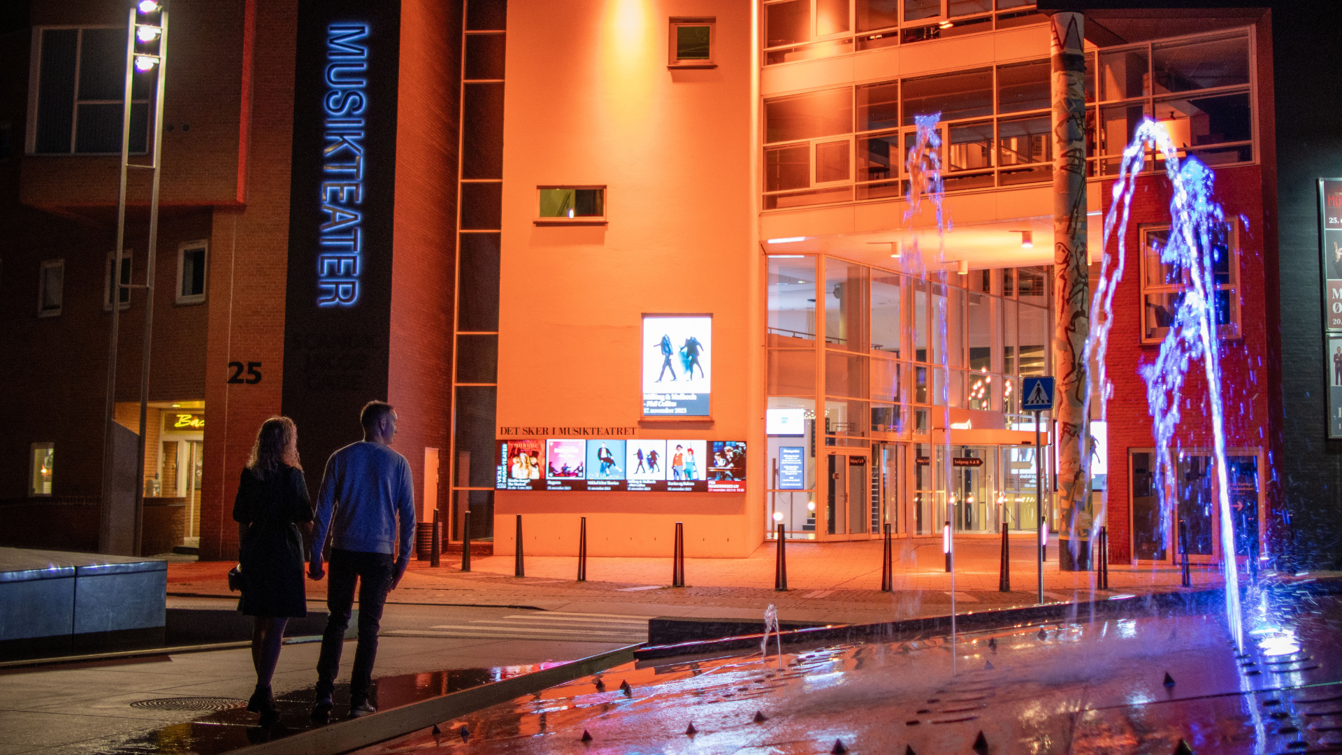 Ein Paar geht auf dem Weg zum Vejle Musikteater über den Kreisverkehr mit dem Brunnen vor dem Musiktheater